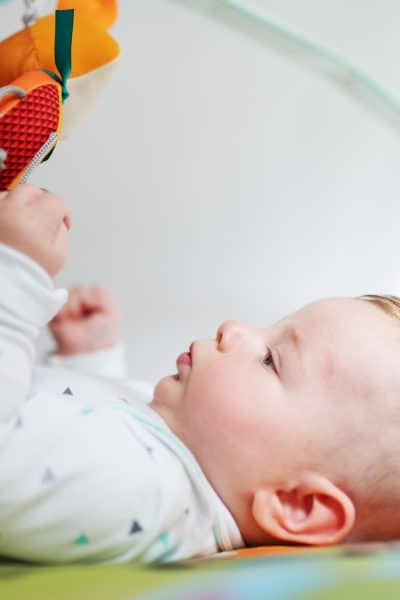 Side view of beautiful curious caucasian six months old baby boy lying in bed and playing with non-toxic baby gym toys.