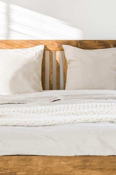 Close-up of double wooden bed with bedding, pillows and blanket against white wall in a bright sunny bedroom interior. Two green plants standing beside. Panorama. Real photo.